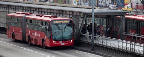Red bus in font of covered pedestrian walkway/station. Partially visible bus on the other side of the station, a few pedestrians visible near the end of the covered walkway.
